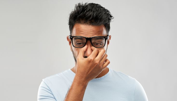 A man wearing glasses, pinching the top of his nose and looking tired.